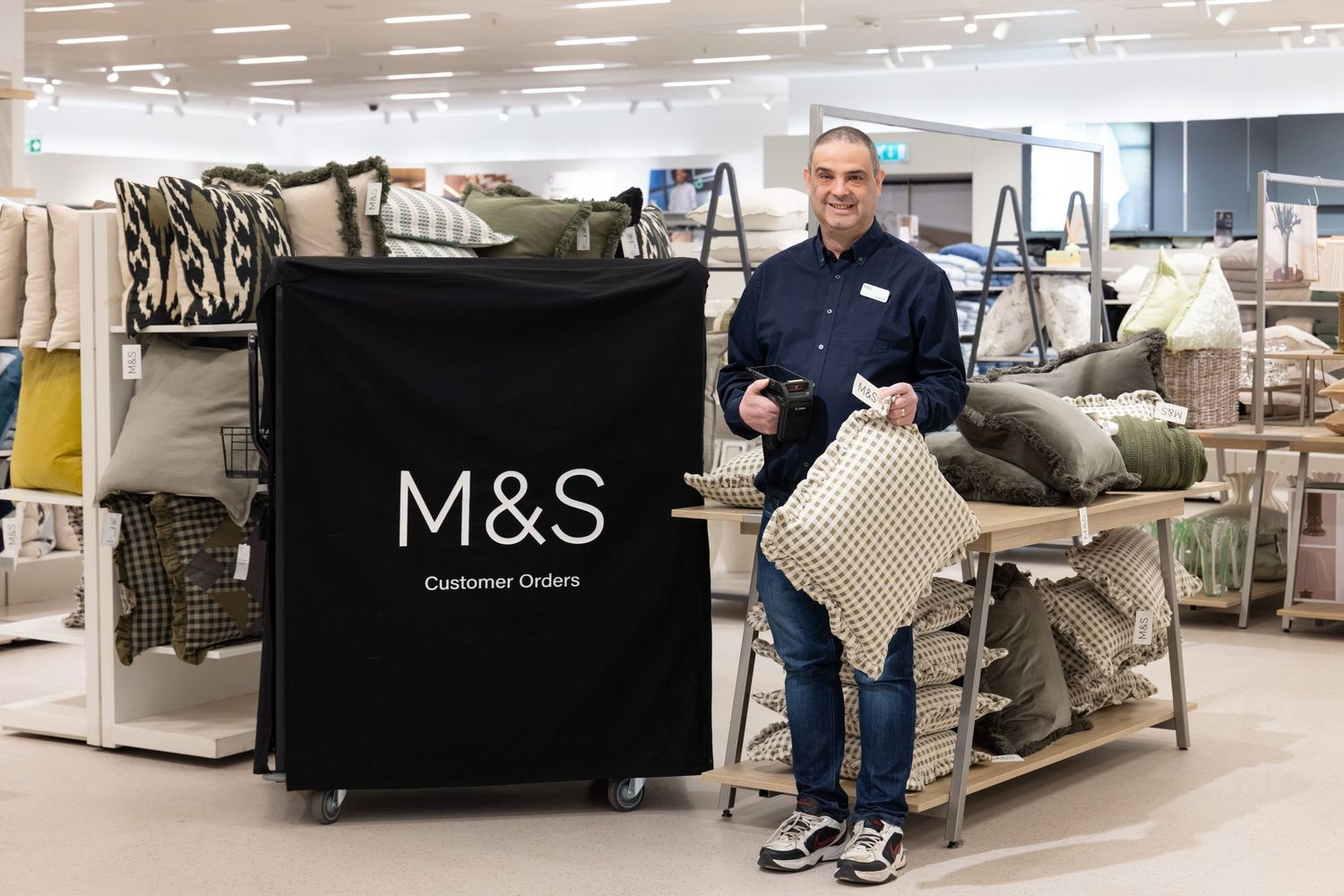 Corporate workplace photography of a Marks & Spencer employee smiling beside a Customer Orders trolley in an M&S home department.