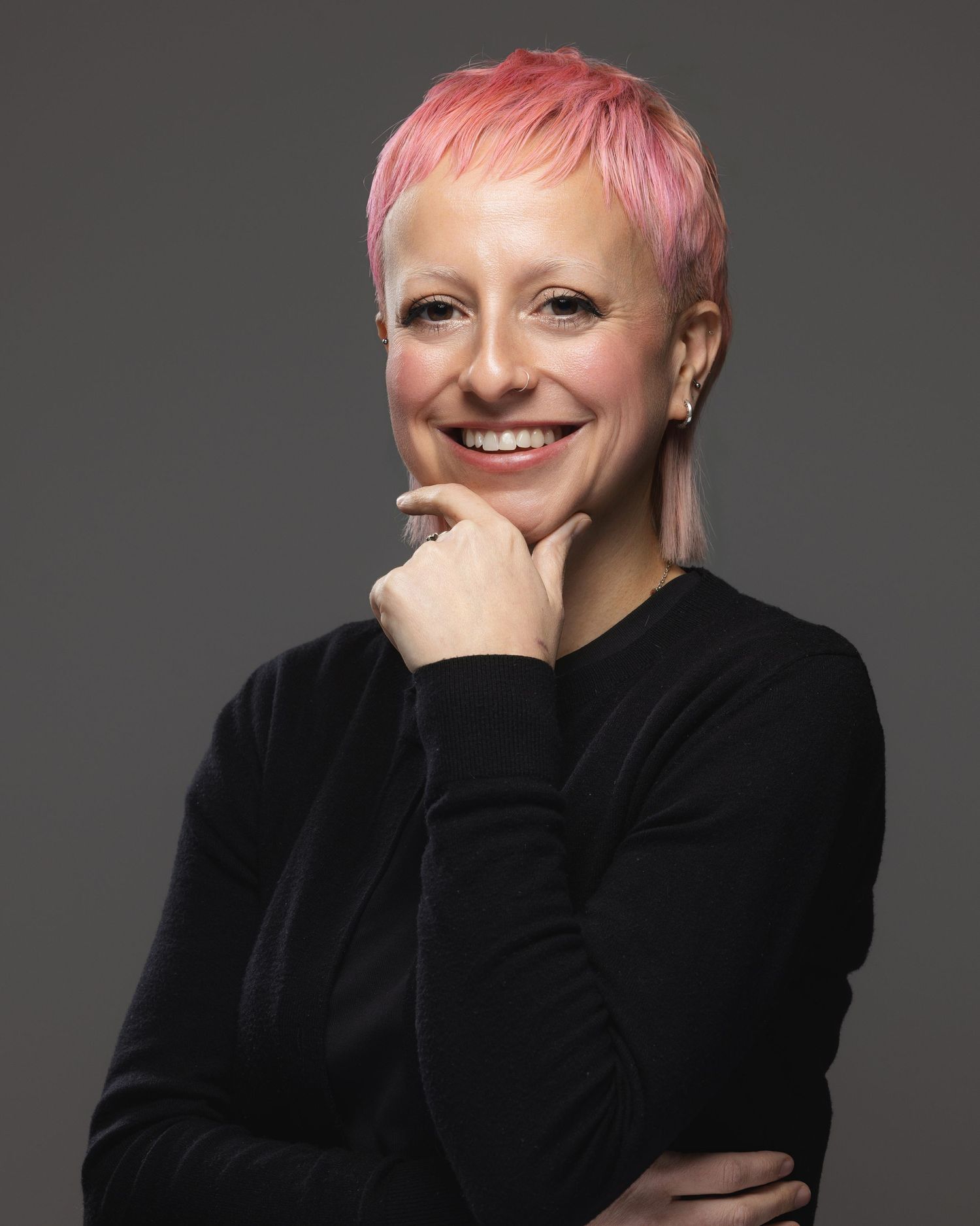 Smiling woman with short pink hair wearing a black sweater, posing confidently against a gray background.