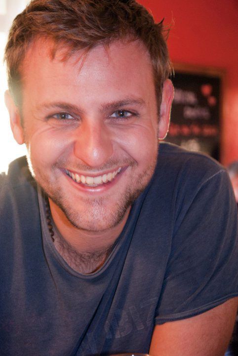 Smiling young man with blue eyes and stubble wearing a navy t-shirt in a warmly lit casual setting.