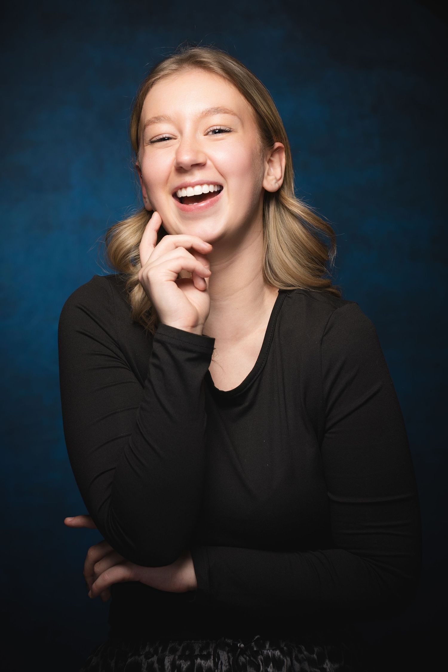Laughing blonde teen girl in black top poses with hand on chin against dark blue studio background.