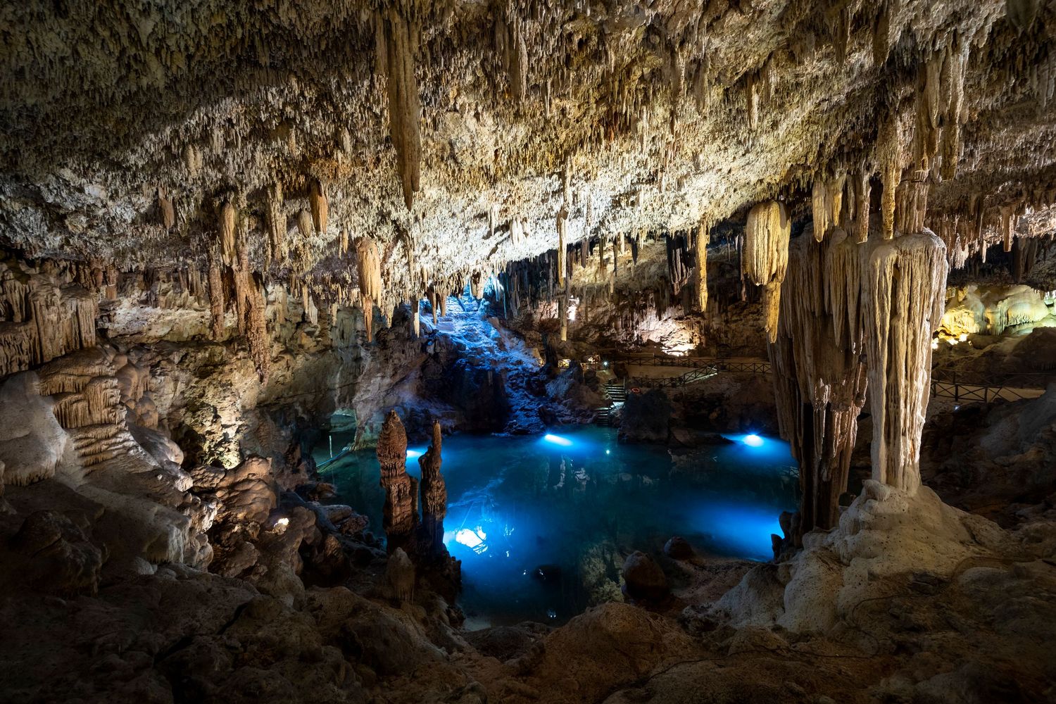 Illuminated underground cave with stalactites and glowing blue cenote pool.