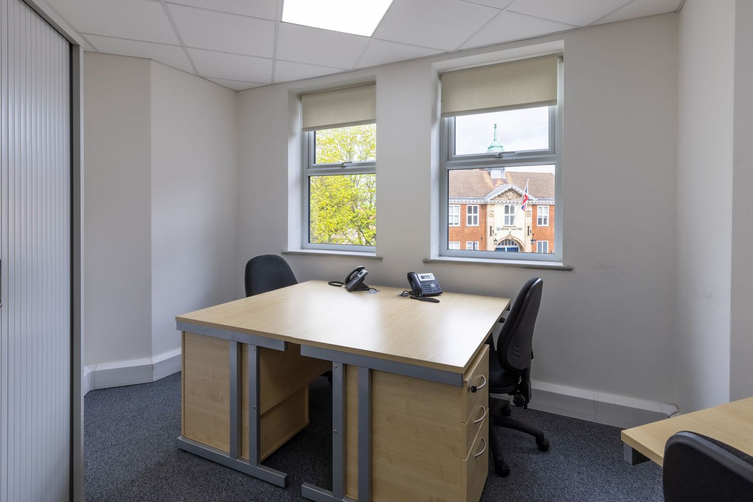Modern small office space with two desks, ergonomic chairs, pedestal drawers, and natural light from two windows.