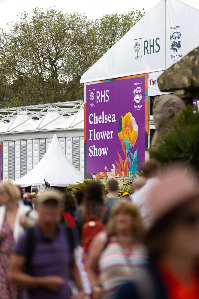 Crowd of visitors gathered at the Chelsea Flower Show with RHS banners and white tents visible in the background.