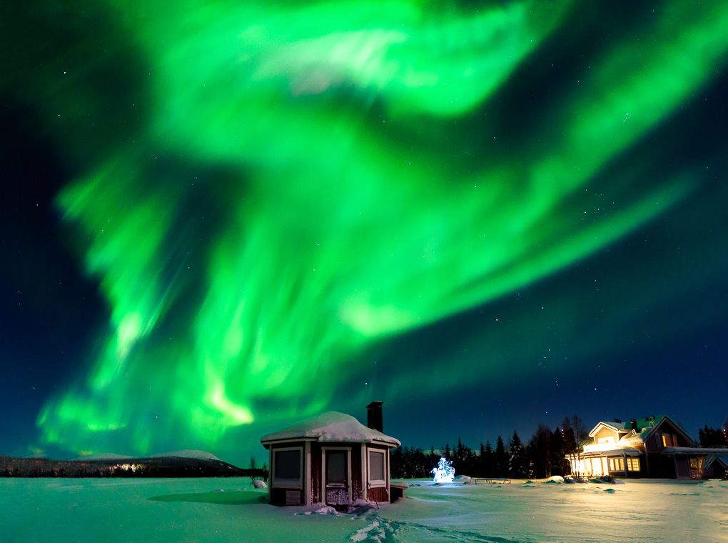 Vibrant green Northern Lights dance across the dark night sky above illuminated buildings in a snowy landscape.