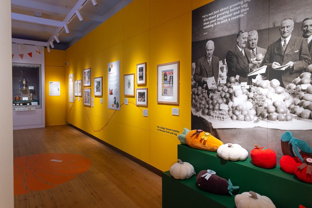 RHS Wisley museum gallery with bright yellow walls, displaying black-and-white photographs and colourful knit items.