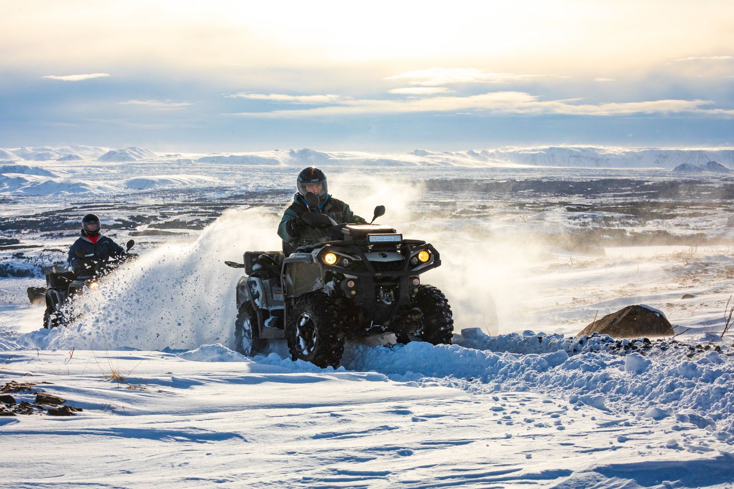 Two ATV riders racing through snowy terrain at sunset with mountains and cloudy skies in the background.