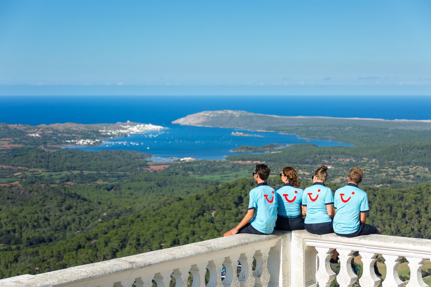 Four people in matching blue shirts sit on a balcony overlooking a scenic coastal bay and forested landscape.