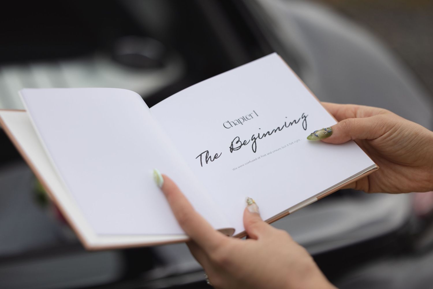 Hands holding open a book to Chapter 1 titled The Beginning, with a car blurred in the background.