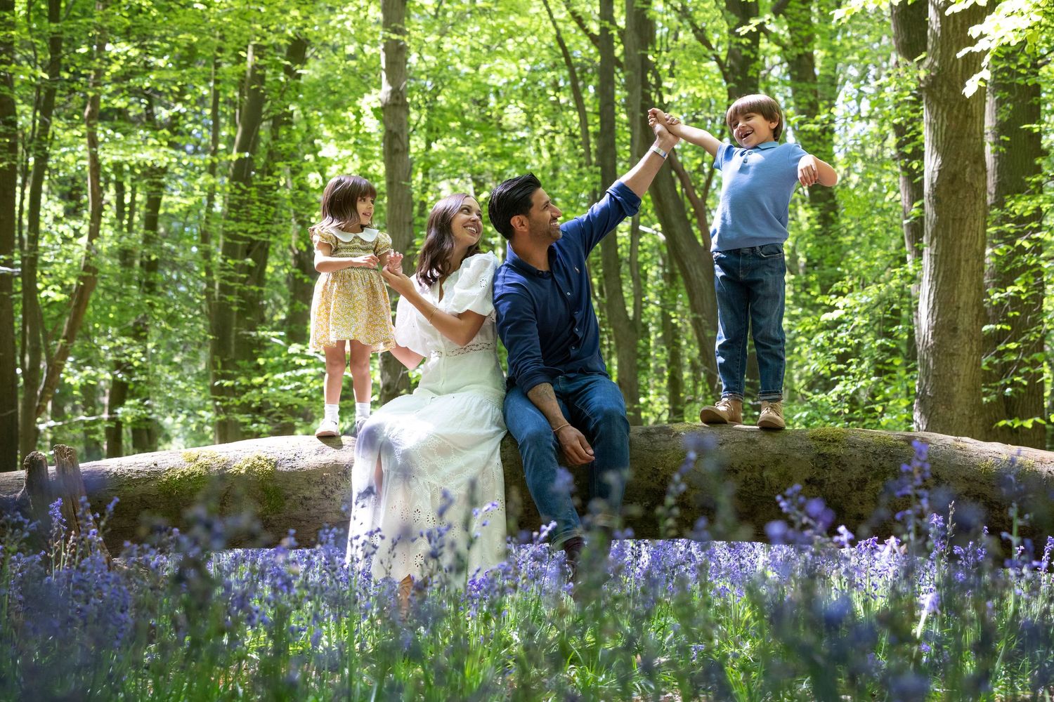 Family enjoying a magical bluebell woodland, sitting on mossy rocks surrounded by purple flowers and lush green trees.