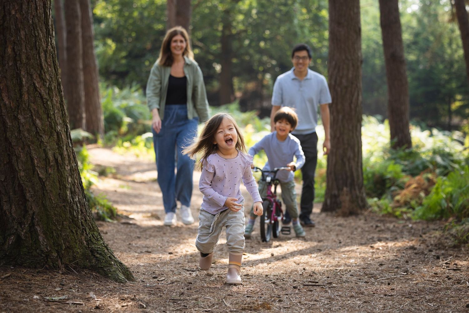 Family enjoying a sunny forest walk, with children playing and parents strolling along a tree-lined path.
