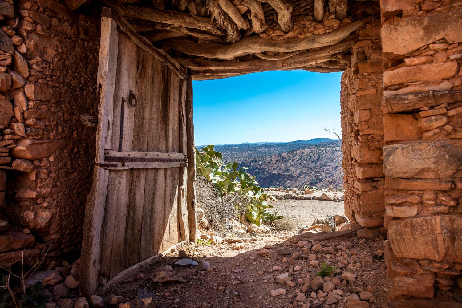 Ancient stone doorway with wooden door opens to a scenic desert canyon vista under a clear blue sky.
