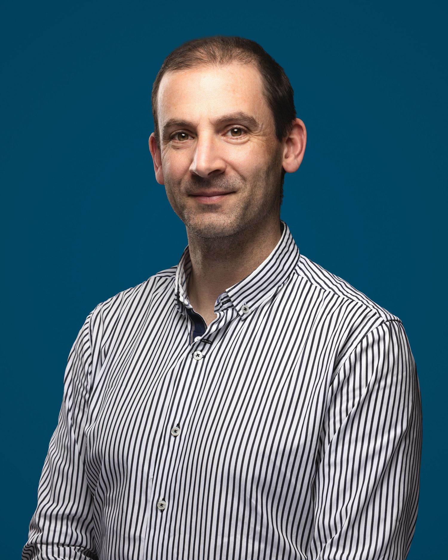 A smiling man in a striped button-up shirt poses against a teal blue background in a professional headshot photo.