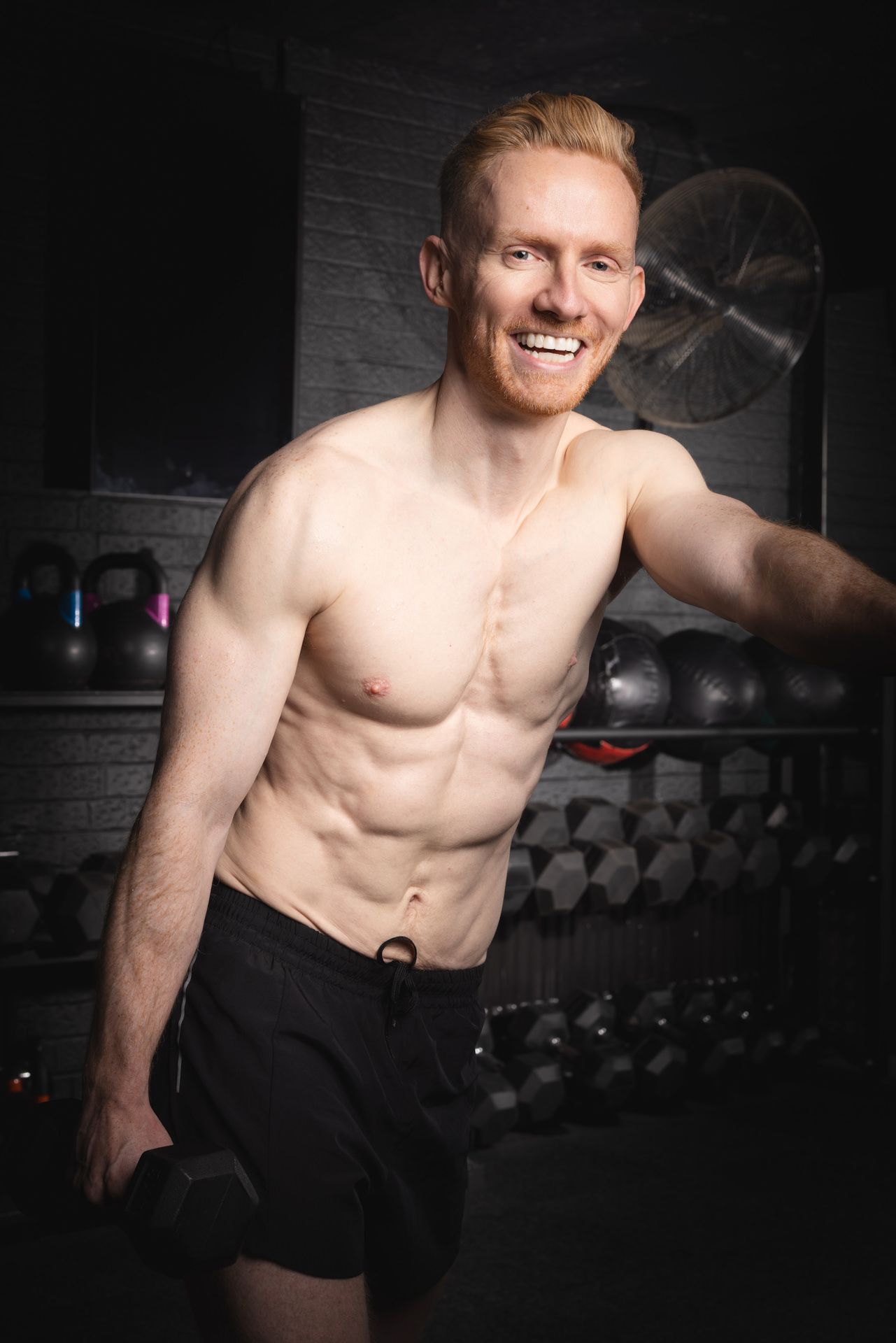 A fitness shoot. A shirtless athletic male in black shorts smiling and posing in a dimly lit Hitchin gym.