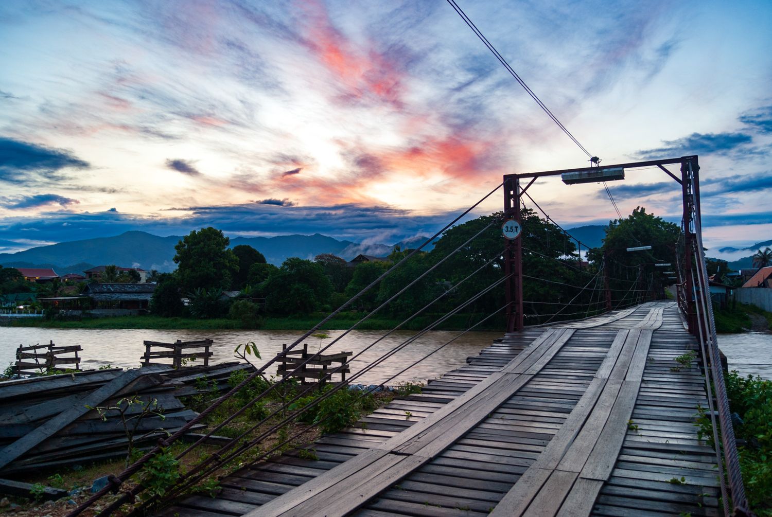 Rustic wooden suspension bridge stretches over river at sunset with dramatic pink and blue sky.