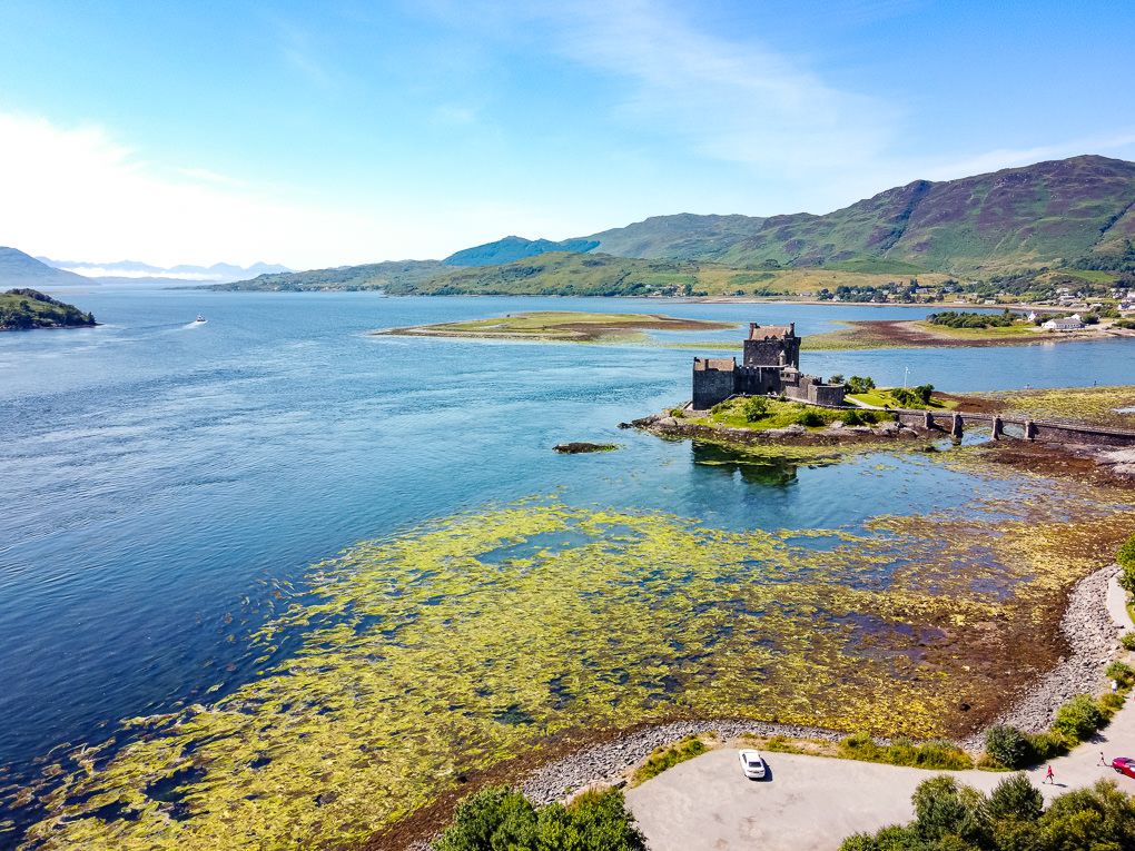Aerial view of Eilean Donan Castle with boats docked along the shoreline and mountains in the background.