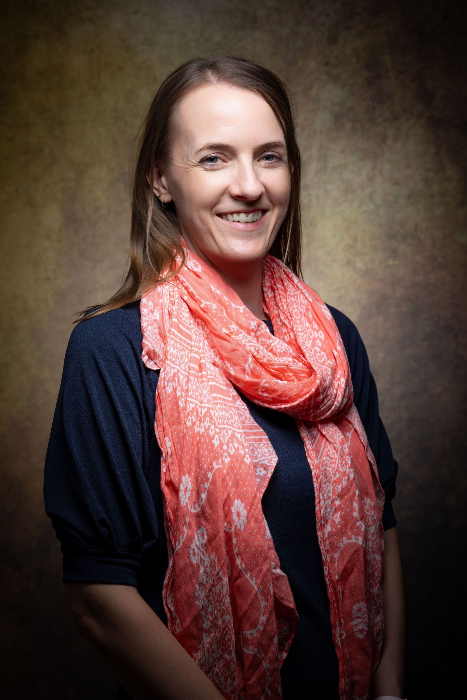 Smiling woman with long hair wearing a navy top and coral floral scarf, posing against a dark textured background.