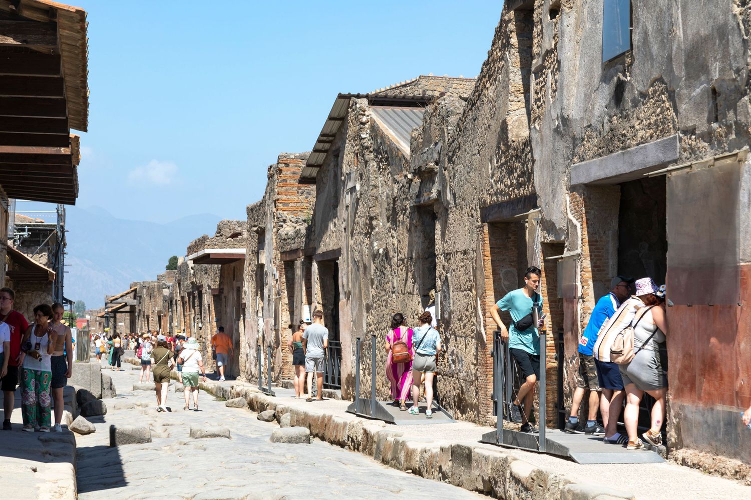 Tourists walk along a cobblestone street lined with ancient stone ruins and weathered doorways in the archaeological site of Pompeii, Italy.