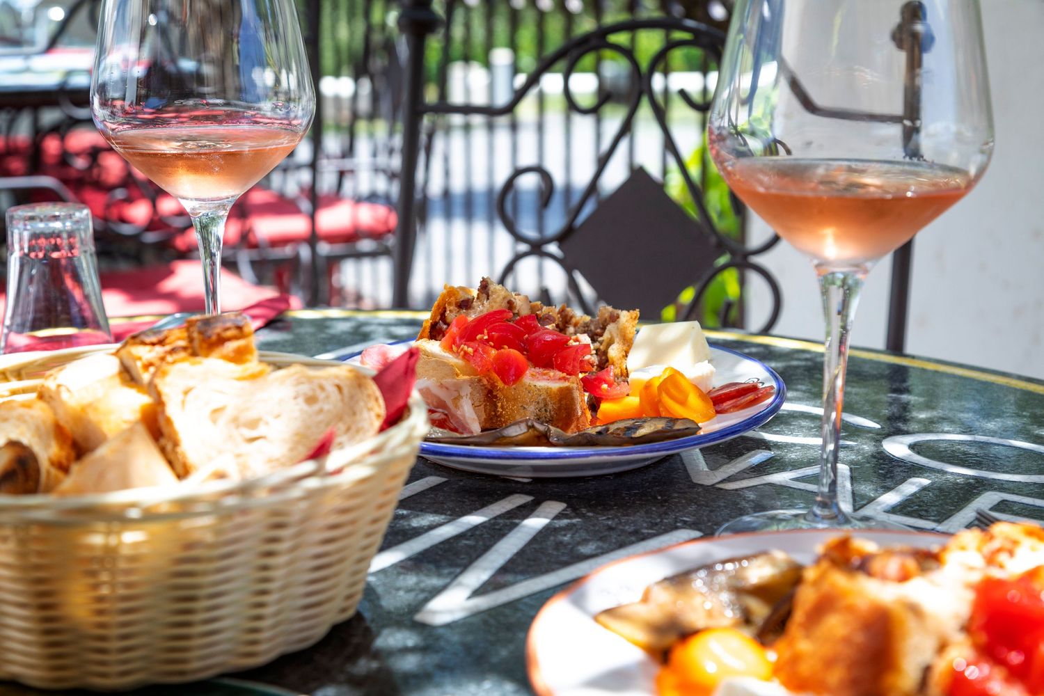 Two glasses of rosé wine sit on an outdoor café table beside a basket of fresh bread and a plate of colorful bruschetta.