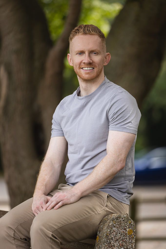 Person in gray t-shirt and khaki pants sits on stone wall outdoors with trees in background.