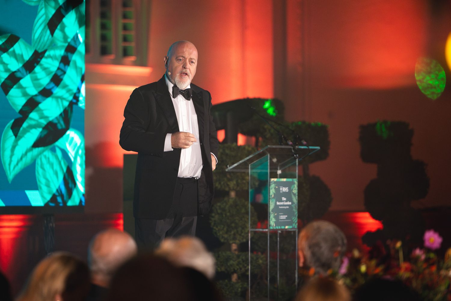 Comedian Bill Bailey, in a black tuxedo, gives a presentation at a formal evening event with red and teal lighting effects.