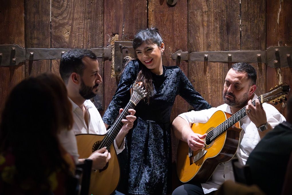 Musicians perform with acoustic guitars while a singer in a dark velvet dress stands between them in a rustic wooden setting.