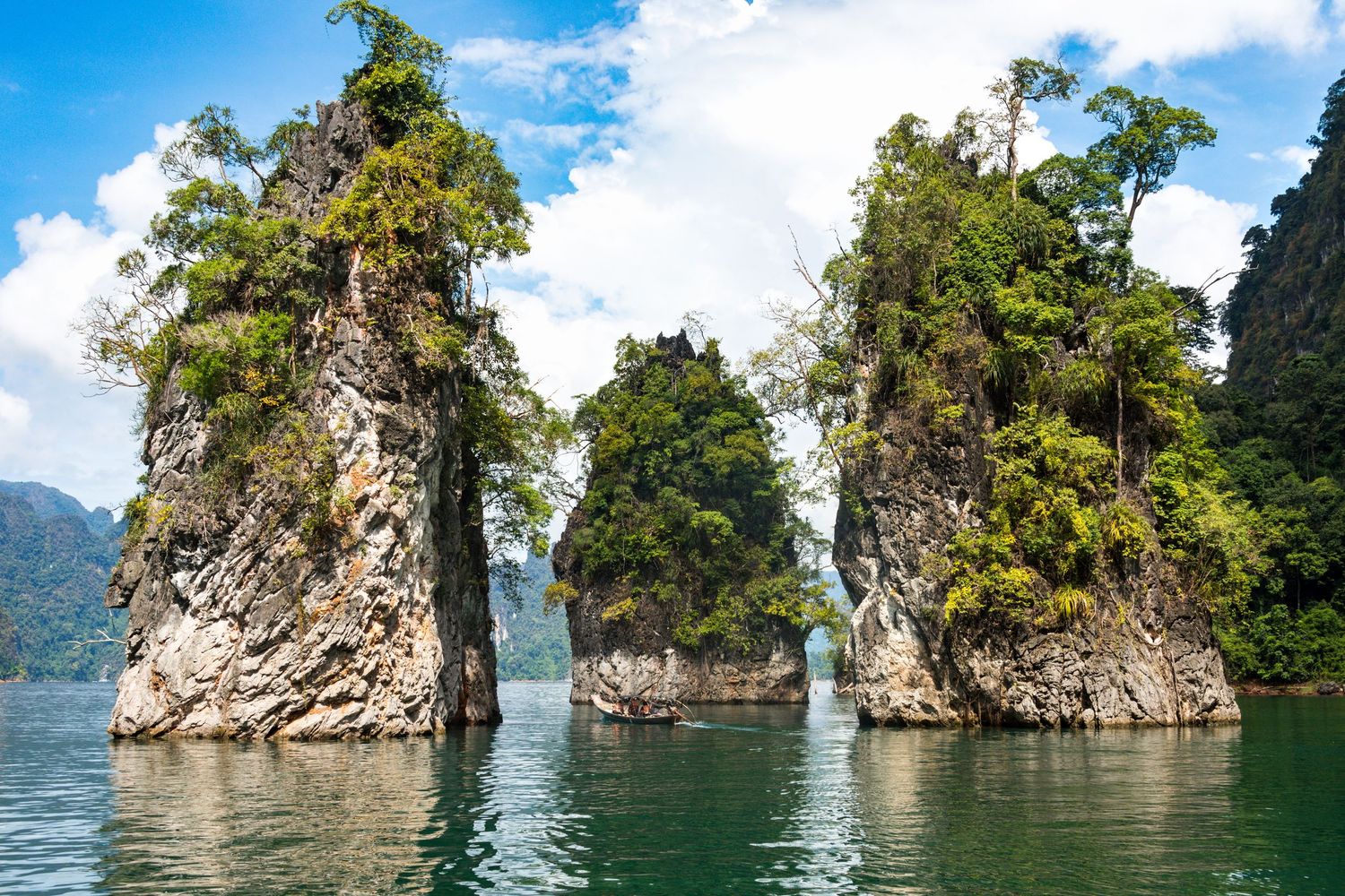 Tall karst limestone rock pillars covered in lush tropical trees rise from a serene green lake in Thailand.