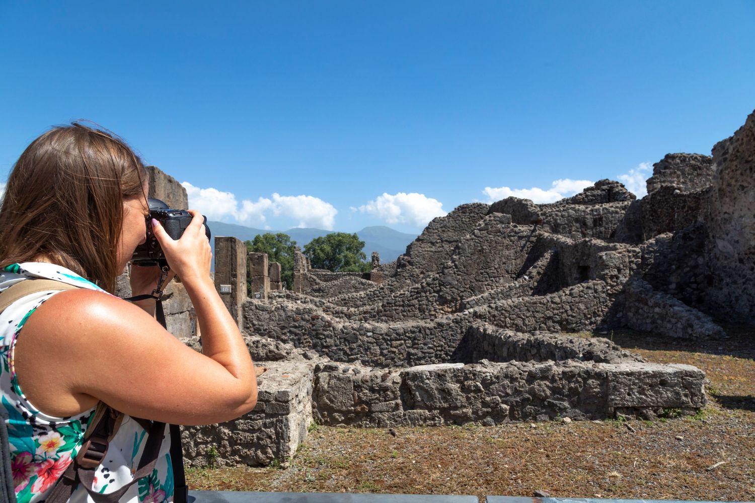 Woman in a floral top photographing ancient stone ruins at Pompeii archaeological site under a bright blue sky with scattered clouds.