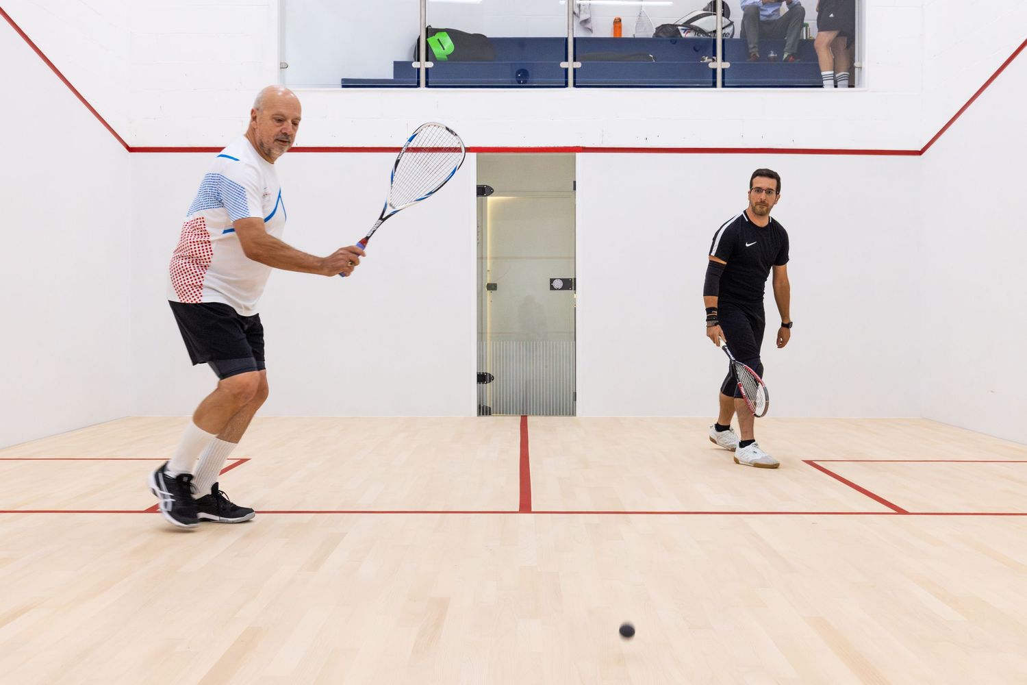 Two men playing squash on an indoor court, one preparing to hit the ball with a racket.