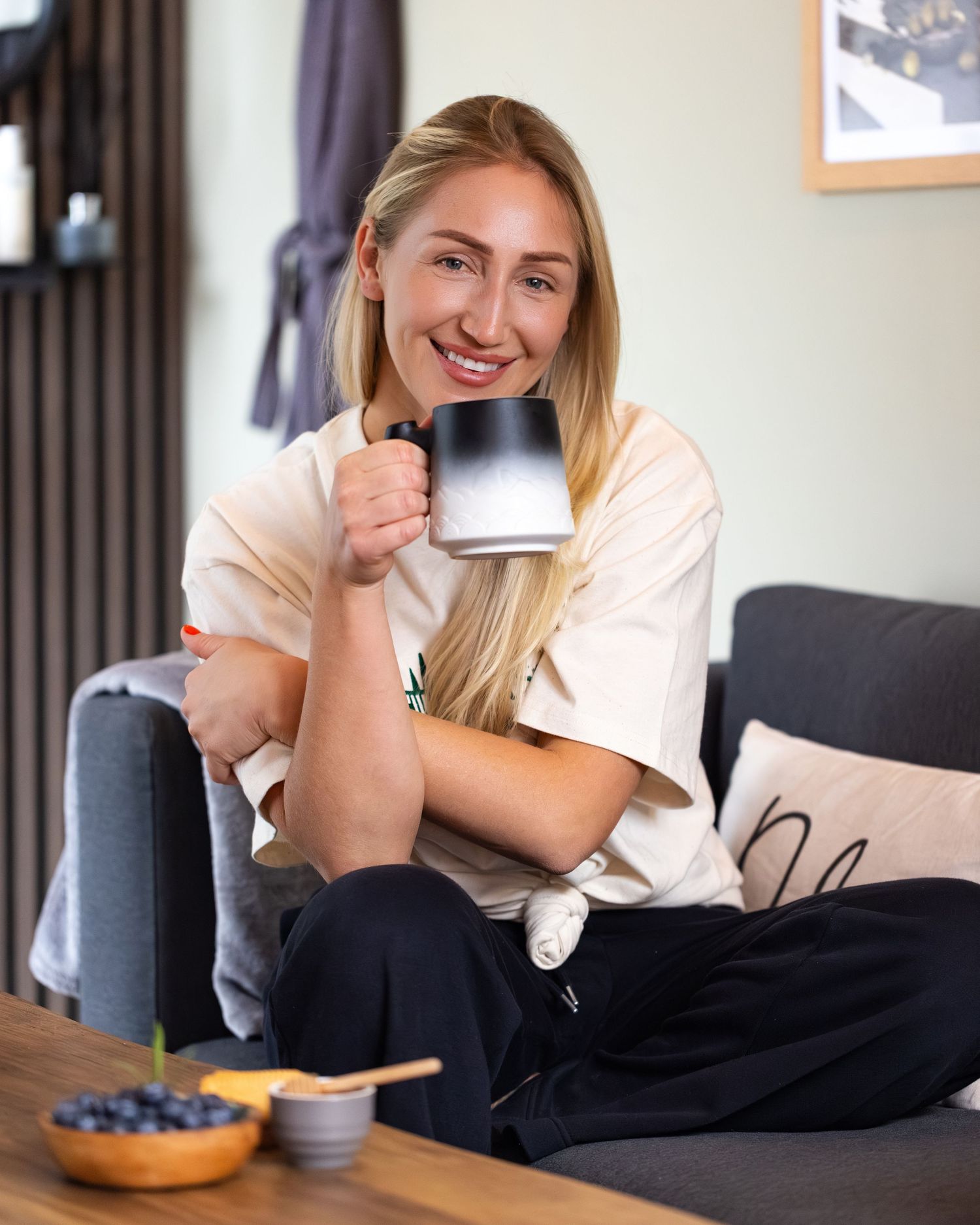 Smiling blonde woman sitting cross-legged on sofa holding black mug with blueberries on wooden table.