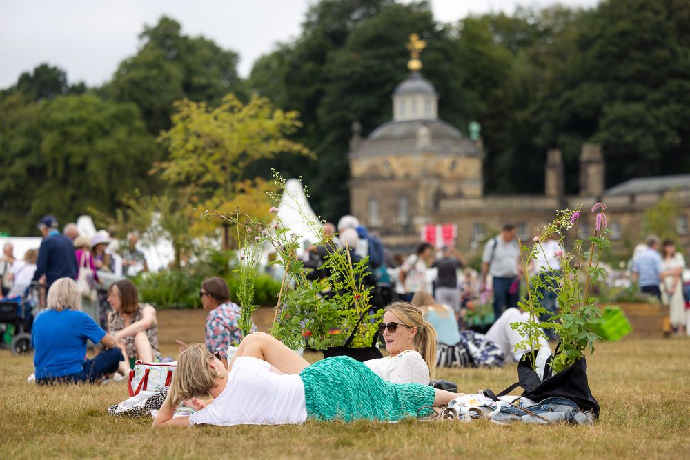 Groups of people enjoying summer picnics on a grassy lawn near ornate historic buildings and gardens.