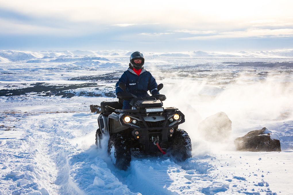A person riding an ATV through snowy terrain with mountains and cloudy skies in the background. Excursion photography in Iceland.
