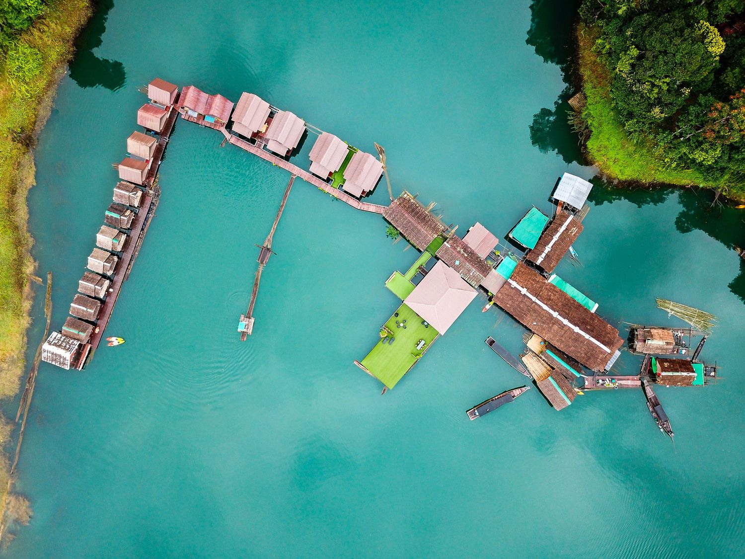 Aerial view of overwater bungalows and floating dock on turquoise water surrounded by lush green trees.