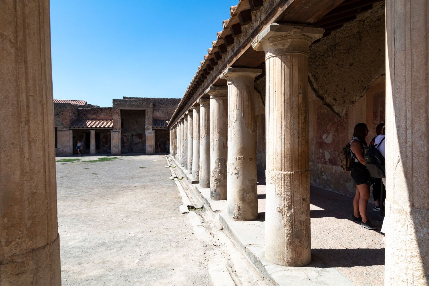 Ancient Roman colonnade with weathered stone columns lining a sunlit courtyard at Pompeii, with visitors standing in the shade.