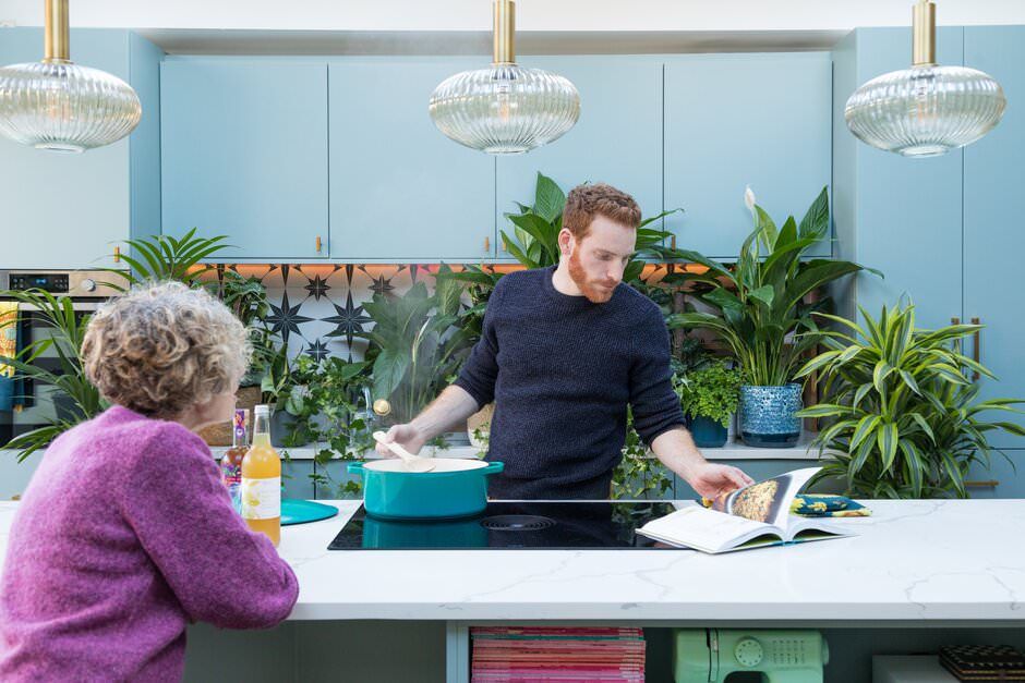 Two people interact in a modern kitchen space with hanging crystal lights and potted plants against blue wall panels.