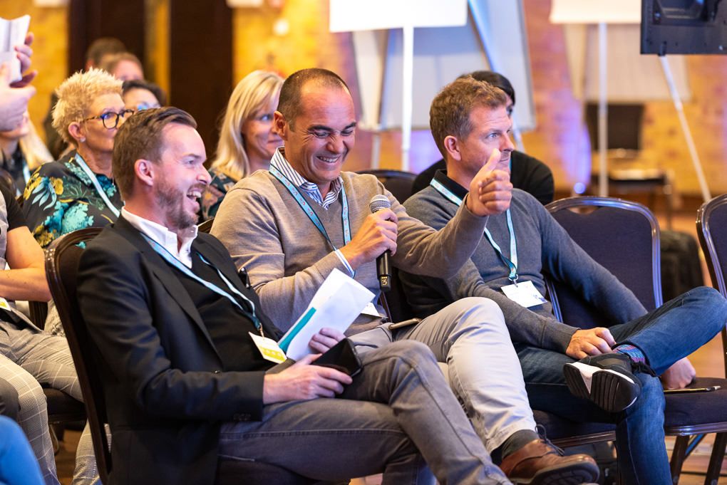 Business professionals seated in rows laugh and smile during a conference presentation or event in London.