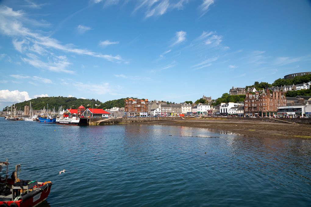 Panoramic view of the harbour in Oban, Scotland, with boats docked along the waterfront and buildings nestled on hillsides.