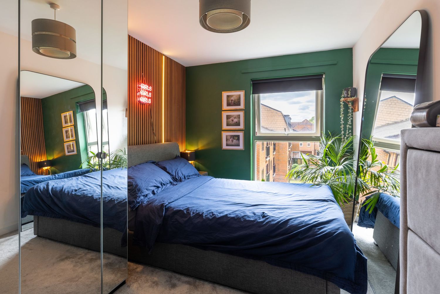 Modern bedroom with green walls, blue bedding, mirrored wardrobe, neon light, and indoor plants near a bright window.