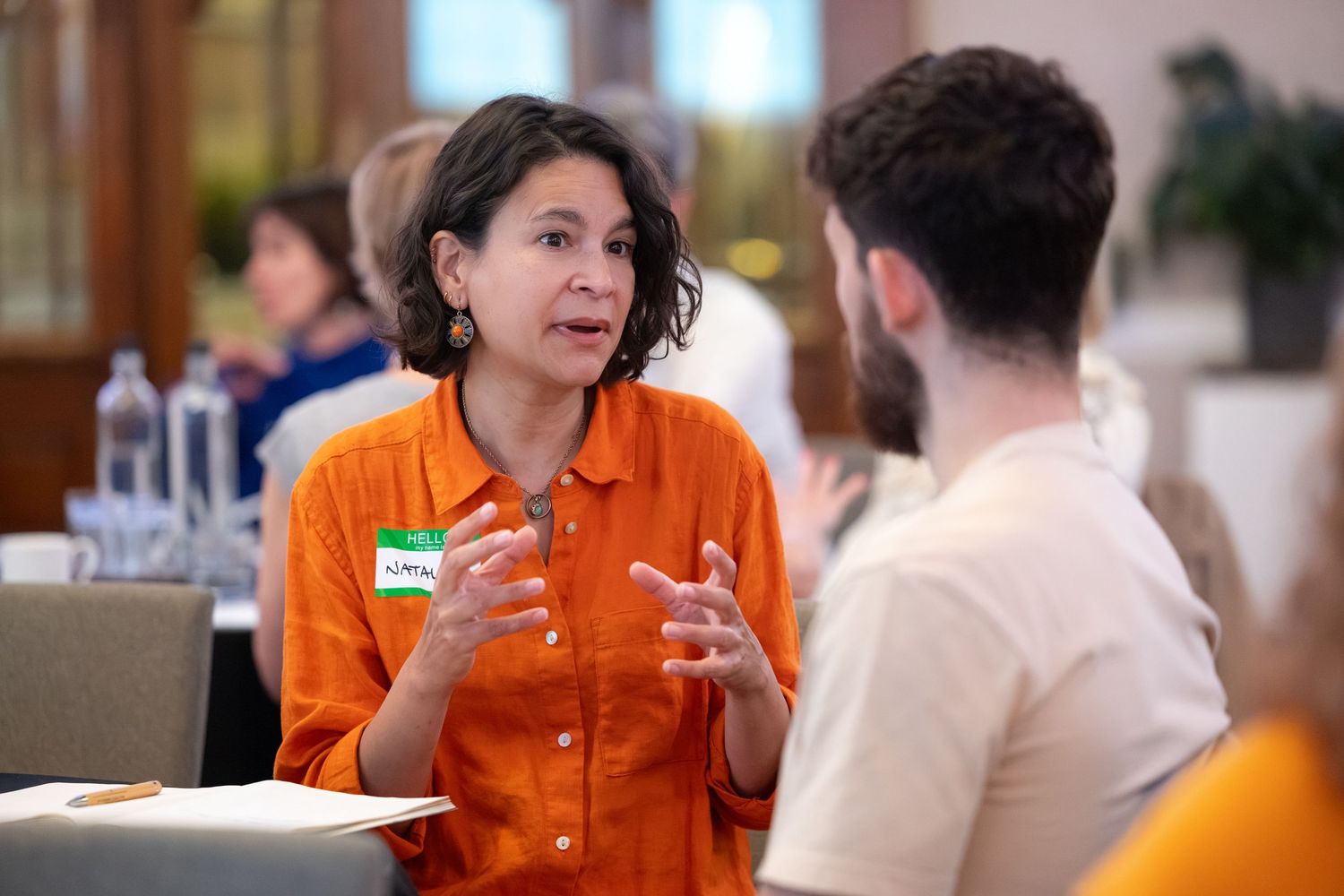 Woman in orange shirt with name tag gestures expressively while speaking to a man at an indoor networking event.