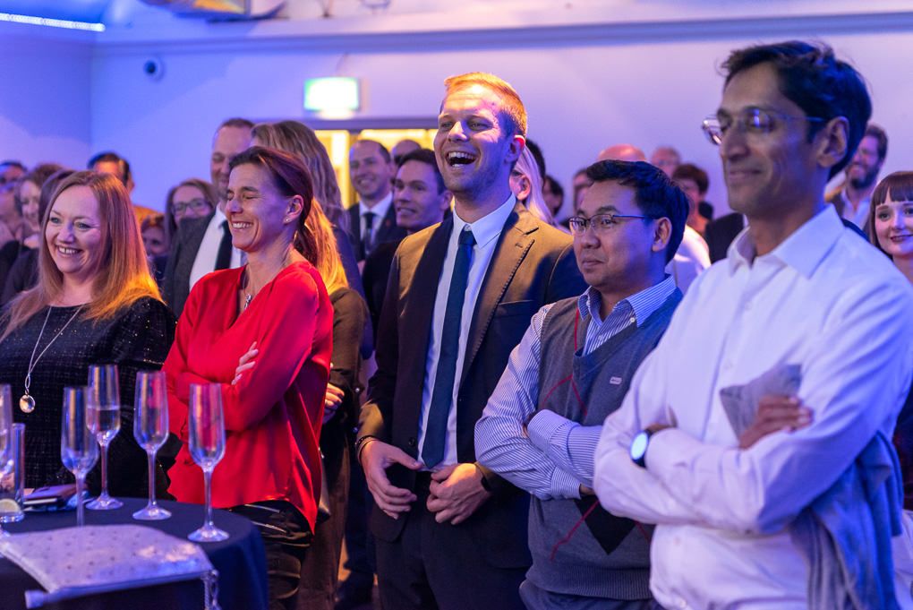 A diverse group of people watching a corporate event together in a room with low blue ambient lighting.
