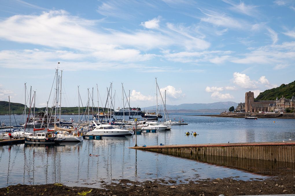 Panoramic view of sailboats moored in the harbour of Oban, Scotland, with blue skies and white clouds overhead.