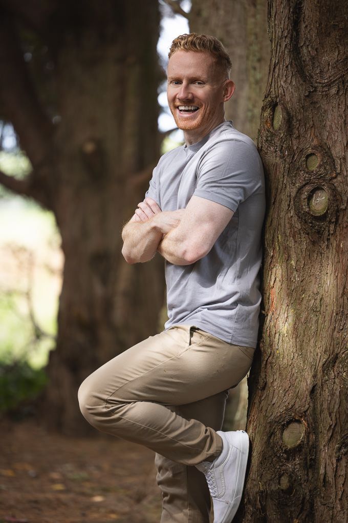 Person wearing a grey t-shirt and khaki pants leans against a large tree trunk in a sunlit forest setting.