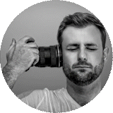 Black and white circular portrait of a bearded man with closed eyes holding a camera with a large lens against his temple.