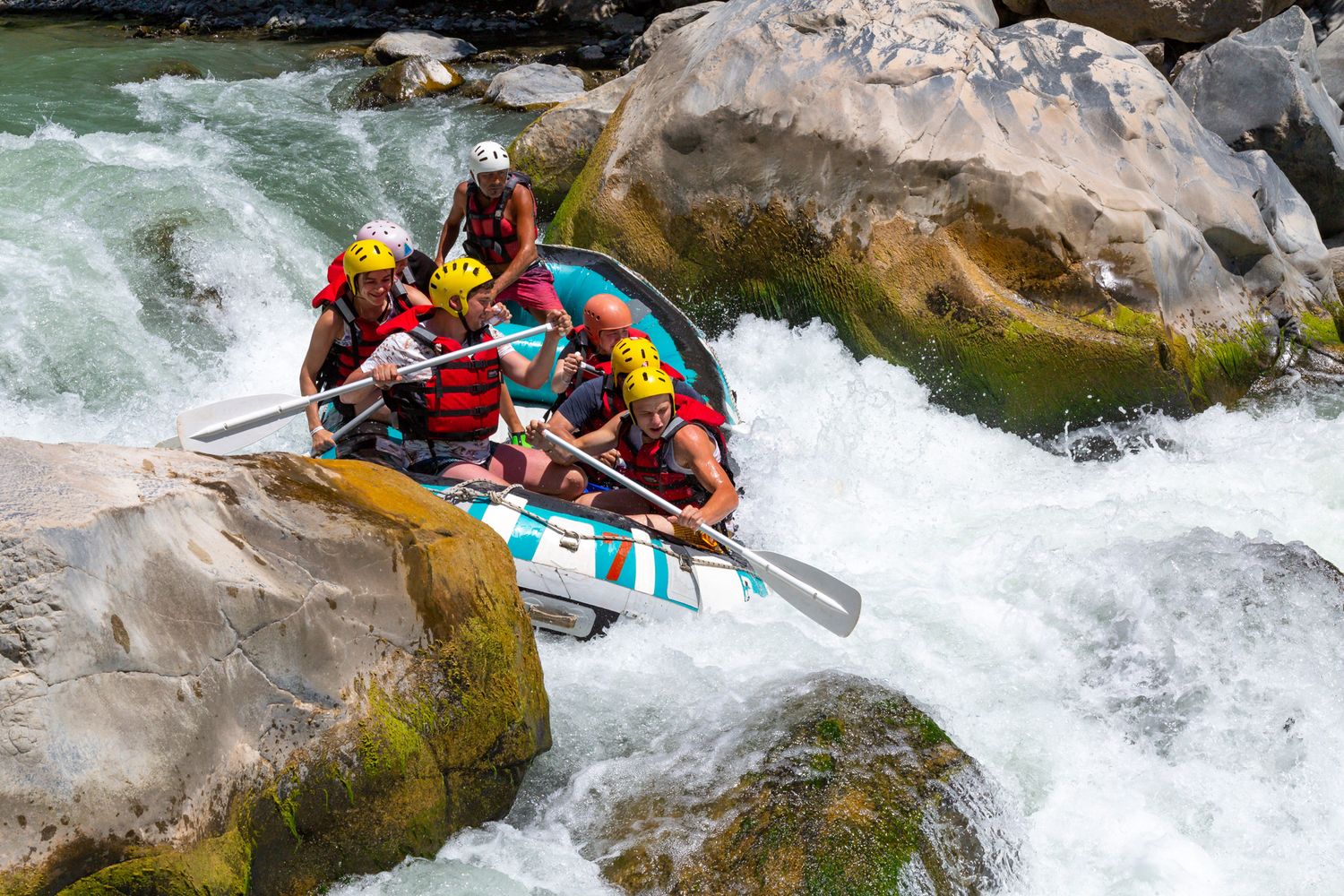 Whitewater rafters in colorful safety gear navigate a turbulent river rapid between large moss-covered boulders.