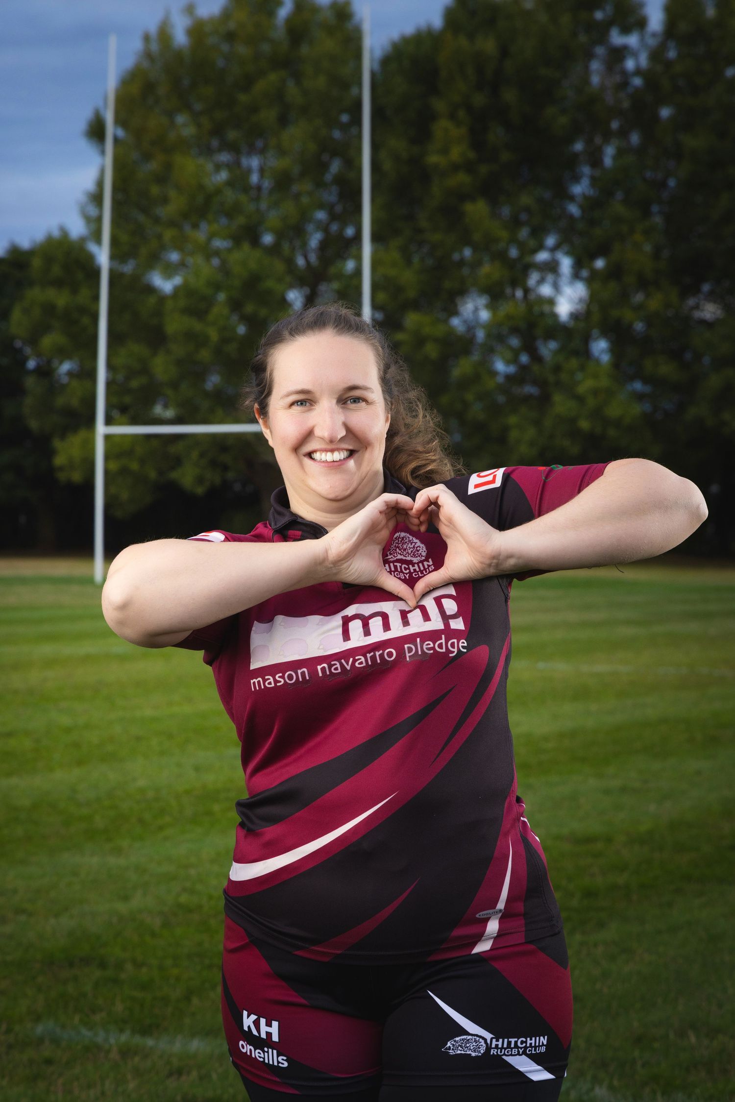 Woman in maroon rugby jersey making a heart shape with her hands on a rugby field with goalposts behind her.
