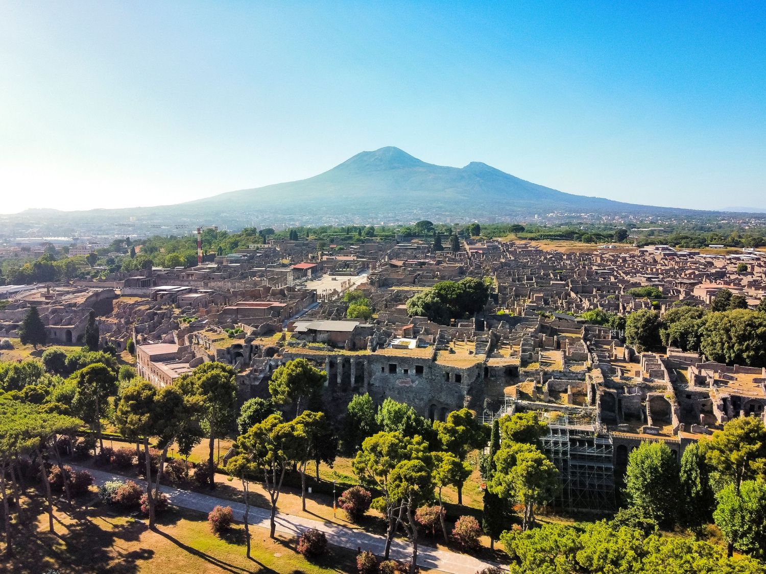 Aerial view of the ancient ruins of Pompeii with Mount Vesuvius rising in the background under a clear blue sky.