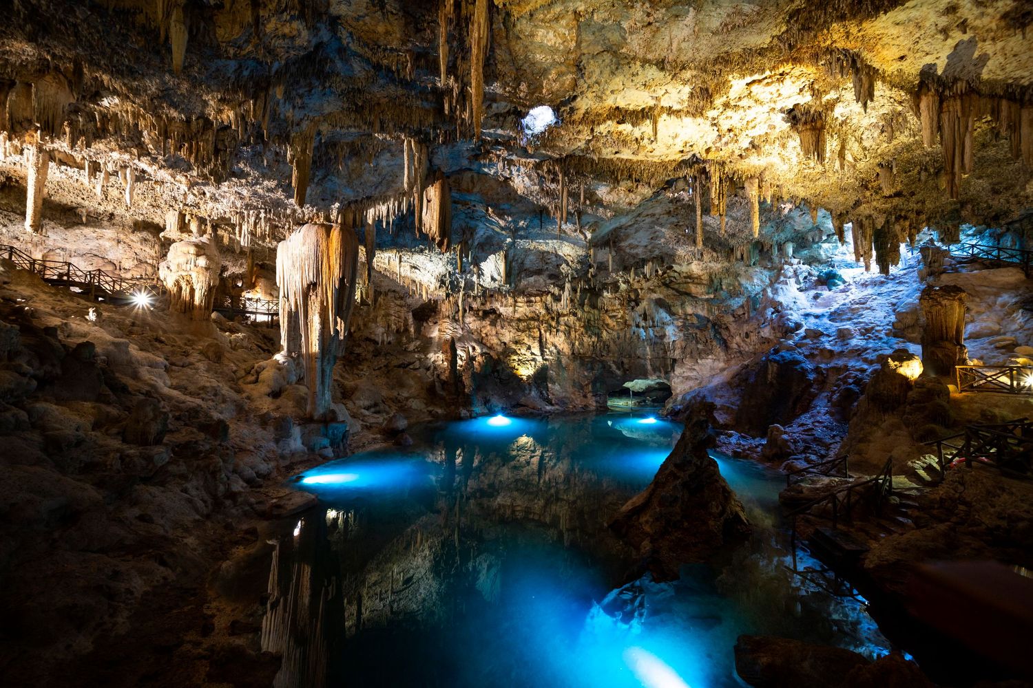 Stunning illuminated cave with stalactites, stalagmites, and glowing blue underground pool reflecting cave formations.
