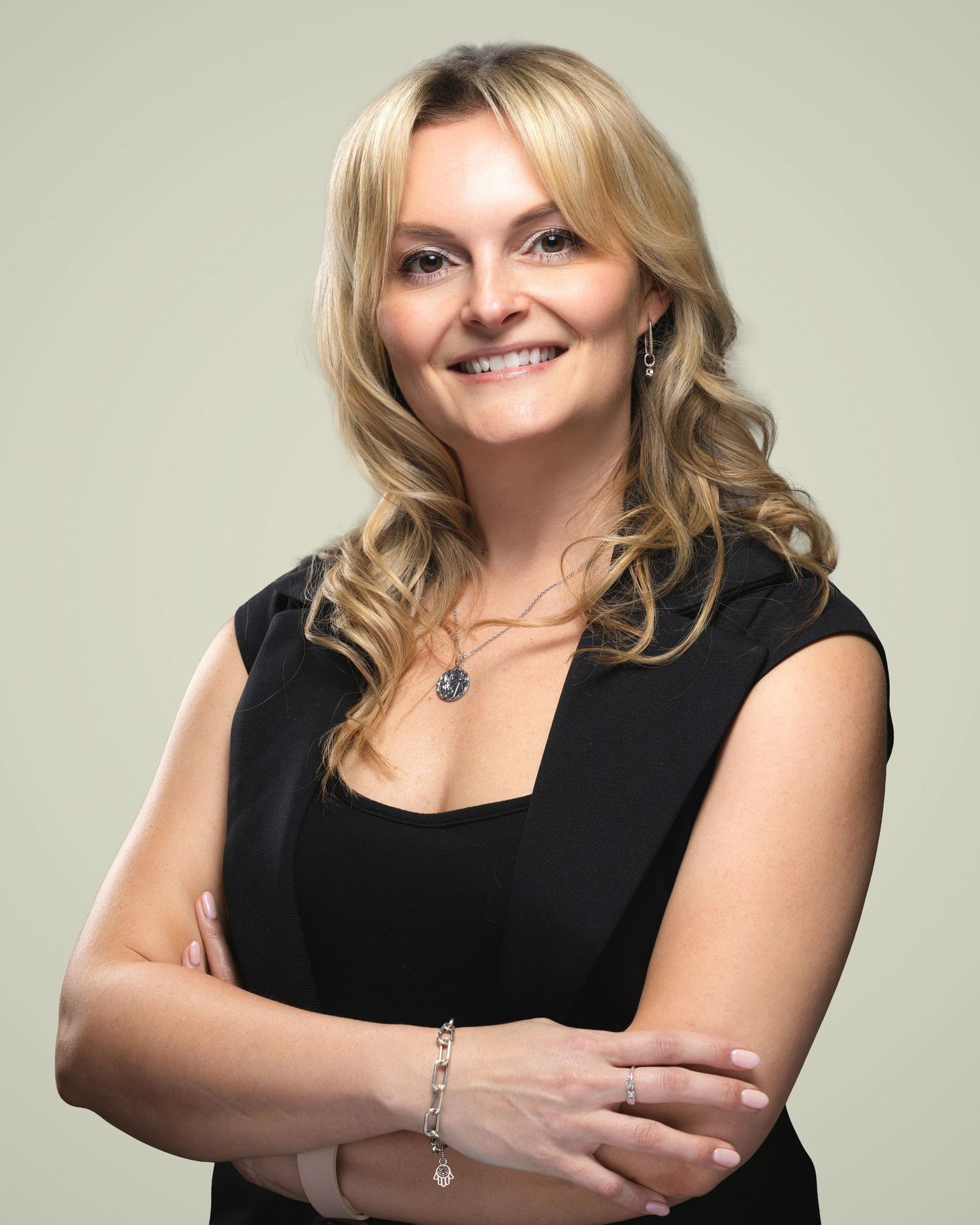 Professional woman with blonde hair in black dress, arms crossed, smiling confidently against neutral background.