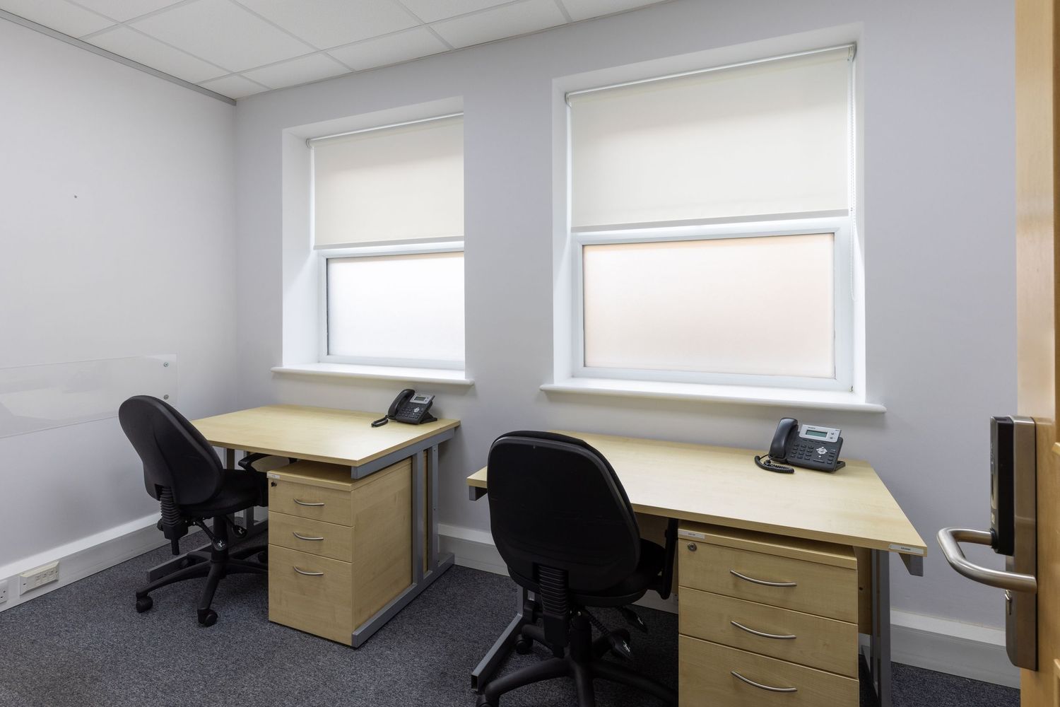 Small office with two wooden desks, black chairs, filing cabinets, desk phones, and two windows with white blinds.