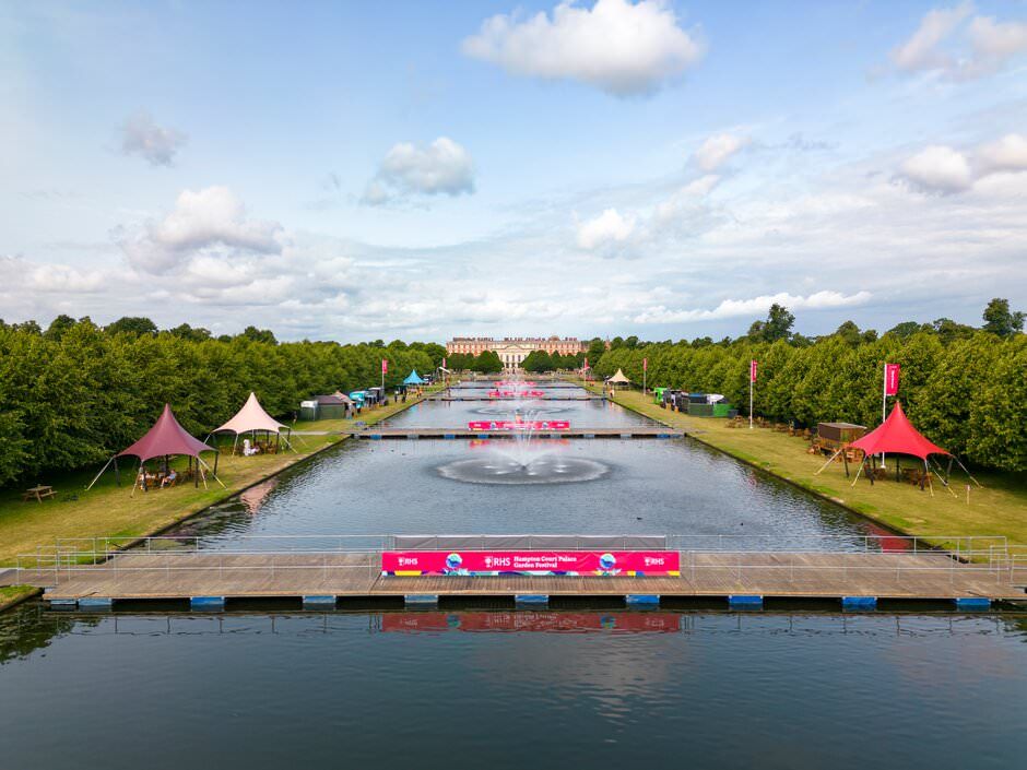 Aerial view of a long waterway with red pontoon boats and covered pavilions along tree-lined banks on a sunny day.
