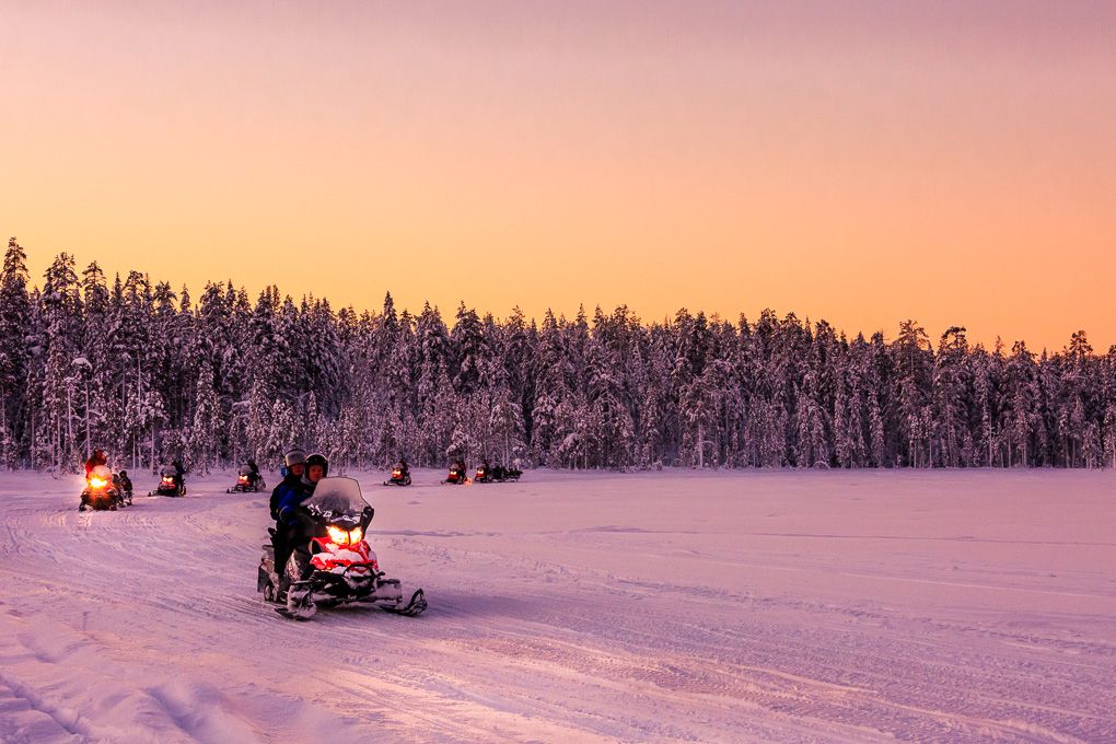 Snowmobiles traverse a snowy landscape at sunset with a snow-covered forest in the background during winter in Lapland. Travel photography.